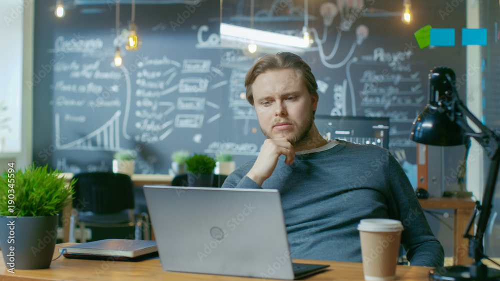 Poster Handsome Young Office Employee Thinks on a Problem Solution While Working on a Laptop Computer. He's Working in the Creative Stylish Office.