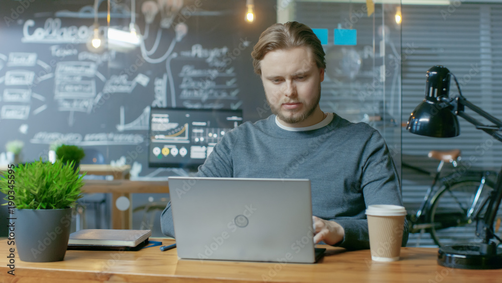 Aufkleber Handsome Young Office Employee Thinks on a Problem Solution While Typing on a Laptop Computer. He's Working in the Creative Stylish Office.