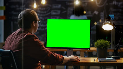 Office Employee at His Desktop Works on a Mock-up Green Screen Personal Computer. Over the Shoulder Footage. His Colleague works in the Background. Creative Office Evening.