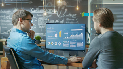 Two Office Employees Have Project Related Discussion, on the Desk Stands Personal Computer with Statistics, Graphs and Charts. They Work in the Creative Office Environment.