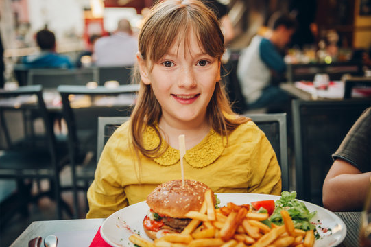 Happy Kid Girl Eating Hamburger And French Fries In Outdoor Restaurant