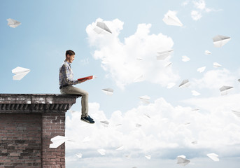 Handsome student guy on roof edge reading book and paper planes fly around