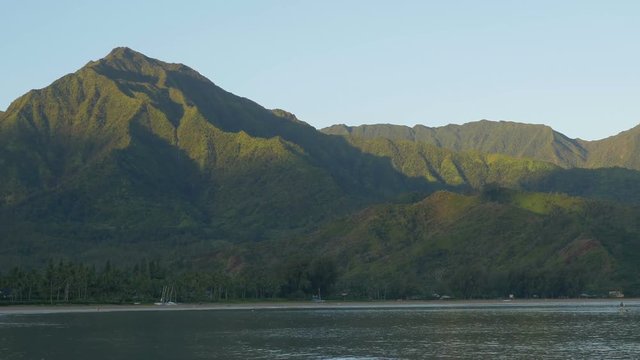Zooming out view of Hanalei Bay, Kauaii, Hawaii, shortly after sunrise, as early morning light strikes surrounding mountain peaks.