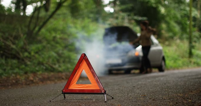 4K Stranded woman with broken down car phoning for assistance on quiet country road. Red warning triangle in foreground. Slow motion.