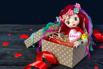 Doll sitting in a gift box with a heart. In the hands of a cookie with the inscription 