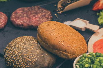 Raw beef burgers, buns with sesame seeds, tomatoes, salad and spices for cooking a hamburger on a dark background