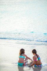Adorable little girls playing with sand on the beach. Back view of kids sitting in shallow water and making a sandcastle
