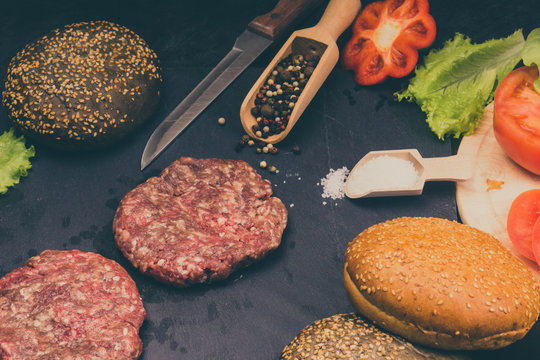 Raw beef burgers, buns with sesame seeds, tomatoes, salad and spices for cooking a hamburger on a dark background