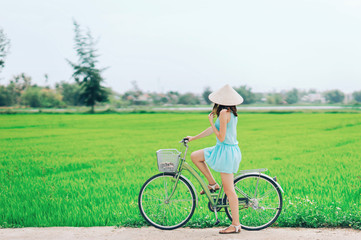 A girl on a bicycle in a traditional Vietnamese hat stands on the road and admires the rice fields.The girl rides a bicycle in a traditional Vietnamese hat in the midst of rice fields, Hoi An, Vietnam