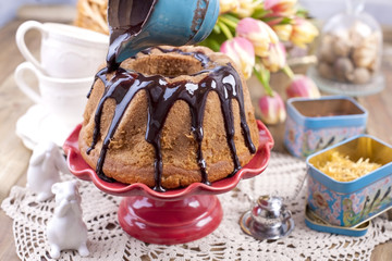 Homemade sweet cake, poured with chocolate, on a red cake plate. On a brown wooden background, dry herbs for tea and a cup. A bouquet of spring flowers. Tulips and a ceramic rabbit. Postcard and free 
