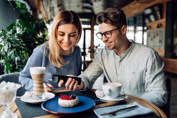 Young smiling guy and girl in headphones, watching a movie on mobile phone while sitting at cafe and drinking coffee.