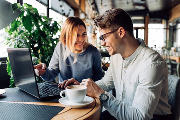 Attractive man in eyeglasses and charming woman is pointing at the laptop screen, laughing together, resting at cafe with cup of coffee.