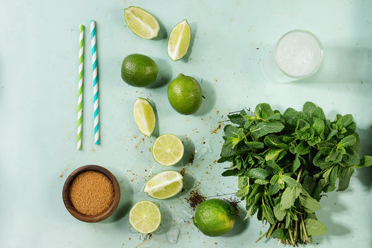 Ingredients for making mojito cocktail. Bundle of fresh mint, limes, brown sugar, crashed ice cubes, glass of soda water, cocktail tubes over green pin up background. Top view, space. Food knolling