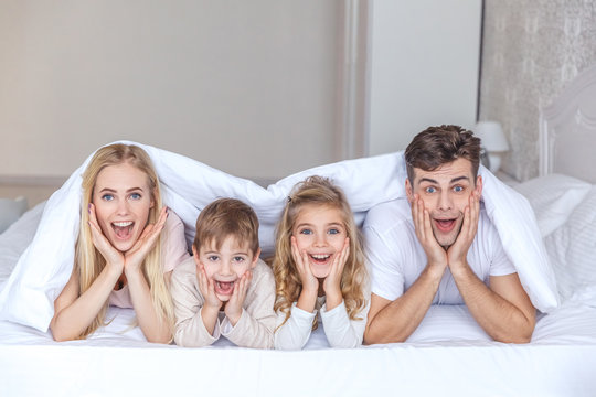 Surprised Young Family Lying In Bed Together Under Blanket And Looking At Camera