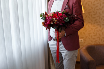 Groom in stylish red suit holding in his hands wedding bridal bouquet of red, white, pink flowers and greenery, with a ribbon of color Marsala
