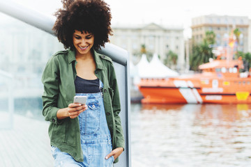 Obraz premium young beautiful dark-skinned girl of mixed race writes an sms message on the smartphone outside.attractive girl downloads a new application on her cellphone against the background of the waterfront
