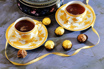Tea ceremony, tea party. Two tea cups of gold color with black tea, candy, chocolate and a box with cookies on a lilac-colored background.