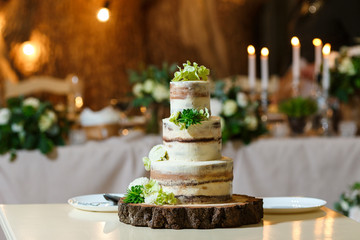 Beautiful multi level naked wedding cake, decorated with cream and fresh flowers, greenery. Delicious dessert at wedding banquet standing on wooden plate