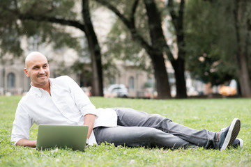 Obraz premium Young contemporary businessman remote working lying down outdoor in a city park using computer - portability, small business, networking concept