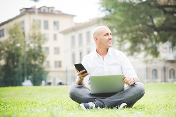 Young contemporary businessman remote working sitting outdoor in a city park using smart phone and computer - portability, small business, networking concept