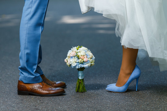 Wedding details: classic brown and blue shoes of bride and groom. Bouquet of roses standing on the ground between them. Newlyweds standing in front of each other