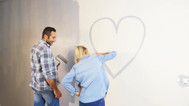 cheerful young love couple in new house painting decorating home and drawing a heart shape on the wall