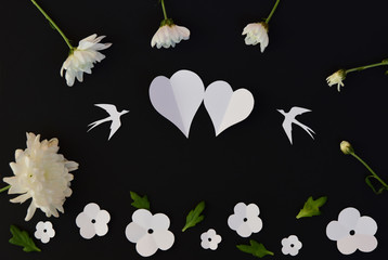 Two paper hearts, two paper birds, white paper and real flowers on black background