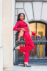 Young African American Woman Fashion in New York. Black Woman dressing in red slim fashionable jacket, pants, black high heel pumps shoes, faux fur collar scarf on shoulder, stands by railing outside