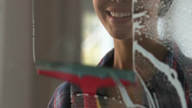 Smiling female cleaner washing glass partitions in office wiping foam and stains