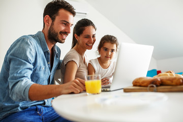 Family breakfast.Young parents with they daughter sitting at the table and watching something on laptop.