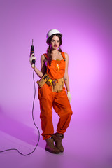 sexy workwoman in overalls and safety helmet with tool belt holding electric drill, on purple