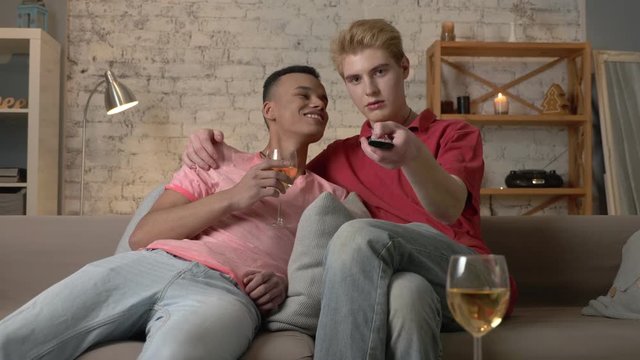 Multinational gay couple sitting on couch, watch TV, use the remote control, look at the camera. Homeliness, romantic evening, hugs, happy LGBT family concept. 60 fps