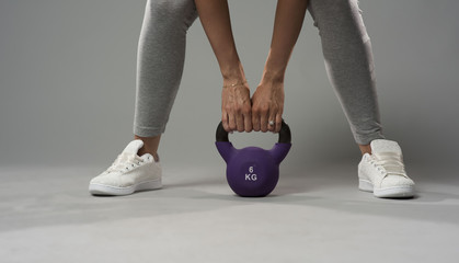 closeup of a woman wearing white sneakers and a grey tights about to lift a purple kettlebell weight from the floor with both hands