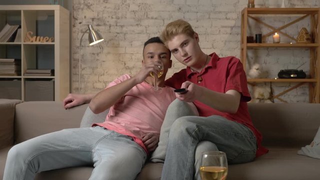 Multinational gay couple sitting on couch, watch TV, use the remote control, look at the camera. Homeliness, romantic evening, cuddles, hugs, happy LGBT family concept. 60 fps