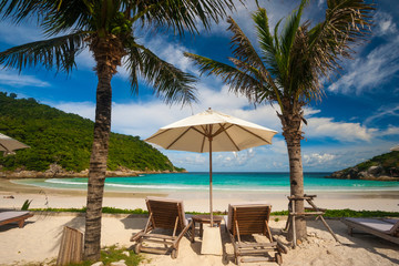 A typical beautiful holiday scene with two beach chairs and an umbrella in between, flanked by palm trees, offering a great view of Patok Beach on Racha Island, Phuket, Thailand.
