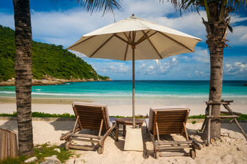 A famous tropical holiday scene with two beach chairs and an umbrella in between, flanked by palm trees, offering a beautiful view of Patok Beach on Racha Island, Phuket, Thailand.