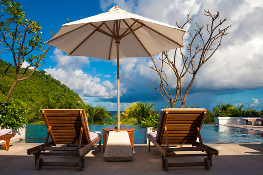 A typical holiday image of two deck chairs next to each other under the shade of a patio umbrella in front of an infinity pool in a tropical paradise.