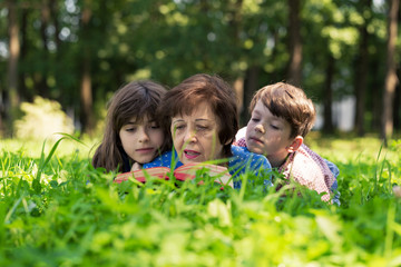 Naklejka na ściany i meble Elderly woman, girl and boy are lying on the lawn and reading a book against green nature background. Grandmother and grandchildren.