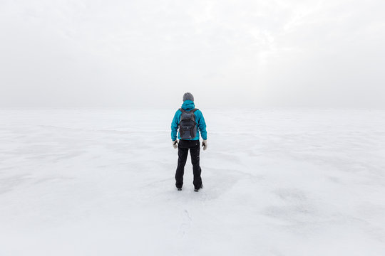 Man with backpack standing alone on the lake ice and staring forward. Cold cloudy winter day