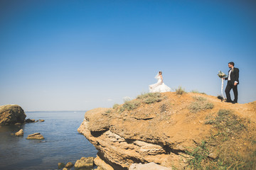 Obraz premium Wedding couple, groom, bride with bouquet posing near sea and blue sky