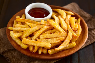 Fresh homemade crispy French fries with a small bowl of ketchup on wooden plate, photographed with natural light (Selective Focus, Focus one third into the fries)