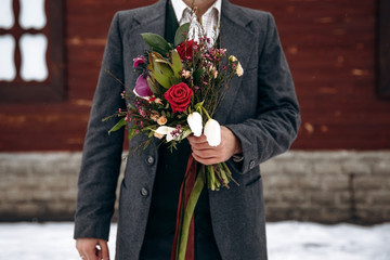 Winter wedding idea. Groom standing in front of wooden house and holding rustic bouquet, is waiting for a bride. Wedding details