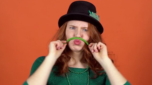 Young woman celebrating saint patrick's day on orange wall in hat grimase