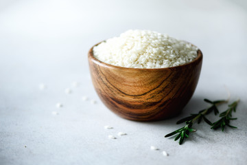 White raw organic jasmine rice in wooden bowl and rosemary on light concrete background. Food ingredients. Copy space