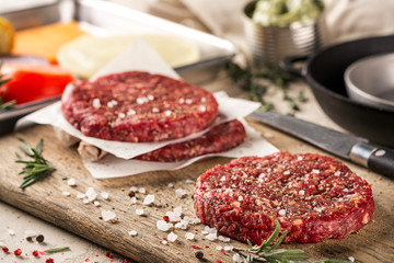 On a wooden cutting board on kraft paper there are raw beef burgers for burgers, spices, rosemary,...