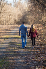 Fototapeta premium Young caucasian couple walking on dim gravel, dirt, rural, country road holding hands, kissing,smiling, laughing in love having fun season late winter bare trees