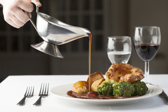 Traditional British roast dinner of rare beef, yorkshire pudding, roast potatoes and brocolli with gravy being poured over from a gravy boat.