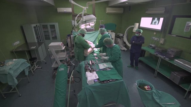 Surgeon orthopedist team with nurse in protective clothes using surgical tools and instruments to operating hip of patient, surgery procedure at operation room, crane shot, wide angle lens, tilt down
