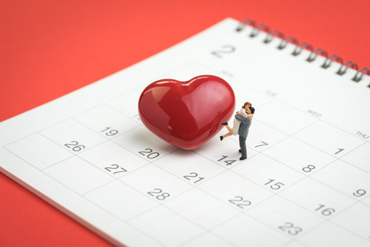 Valentine day concept miniature figures sweet couple standing with shiny red heart shape on 14th February calendar on red background