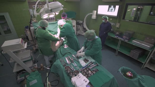 Surgeon team with drill drilling bone during hip replacement, surgery procedure at operation room, patient under anesthesia on table, nurse preparing surgical tool and instruments on tray, crane shot